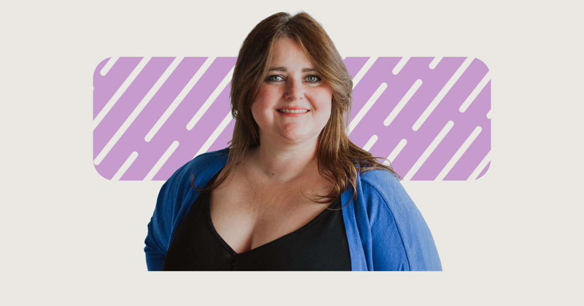 Une femme souriante, vêtue d'un gilet bleu par-dessus un haut noir, se tenant devant un motif décoratif violet.